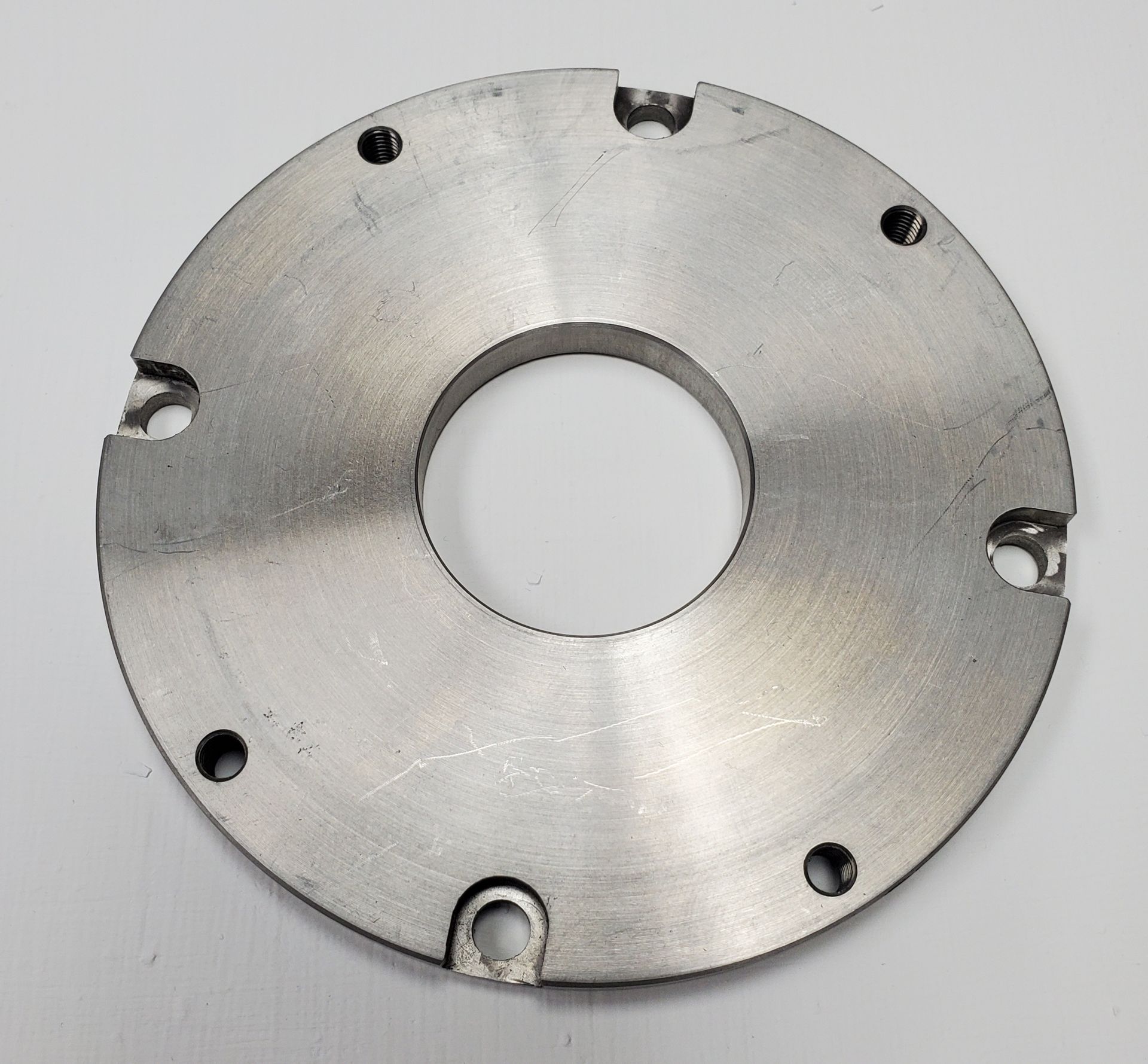 A circular metal mounting plate with a center hole, featuring three edge notches and four smaller pre-drilled holes.