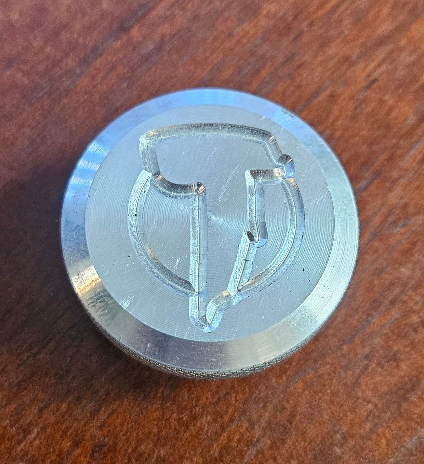 A circular silver metal coin featuring an engraved logo of a stylized letter 'V' within an inverted shield outline.