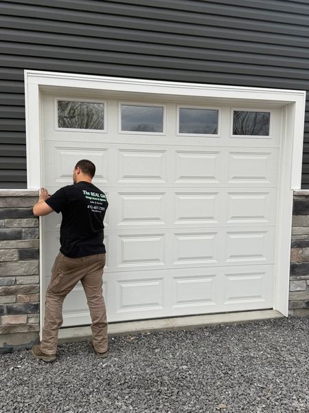 Man installing a large white garage door on a gray house exterior.