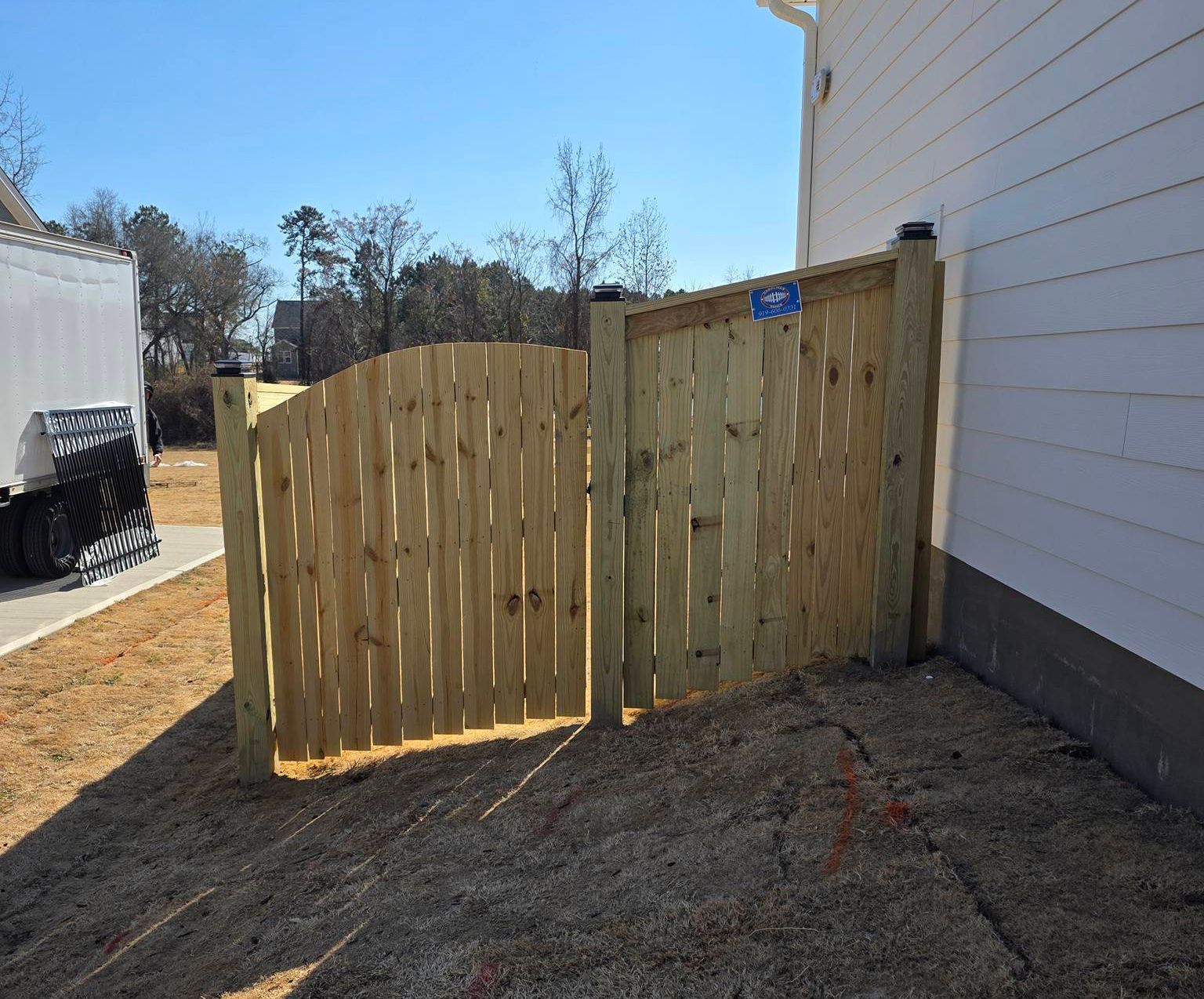 Wooden backyard fence panels beside a white house on a gravel yard under bright sun