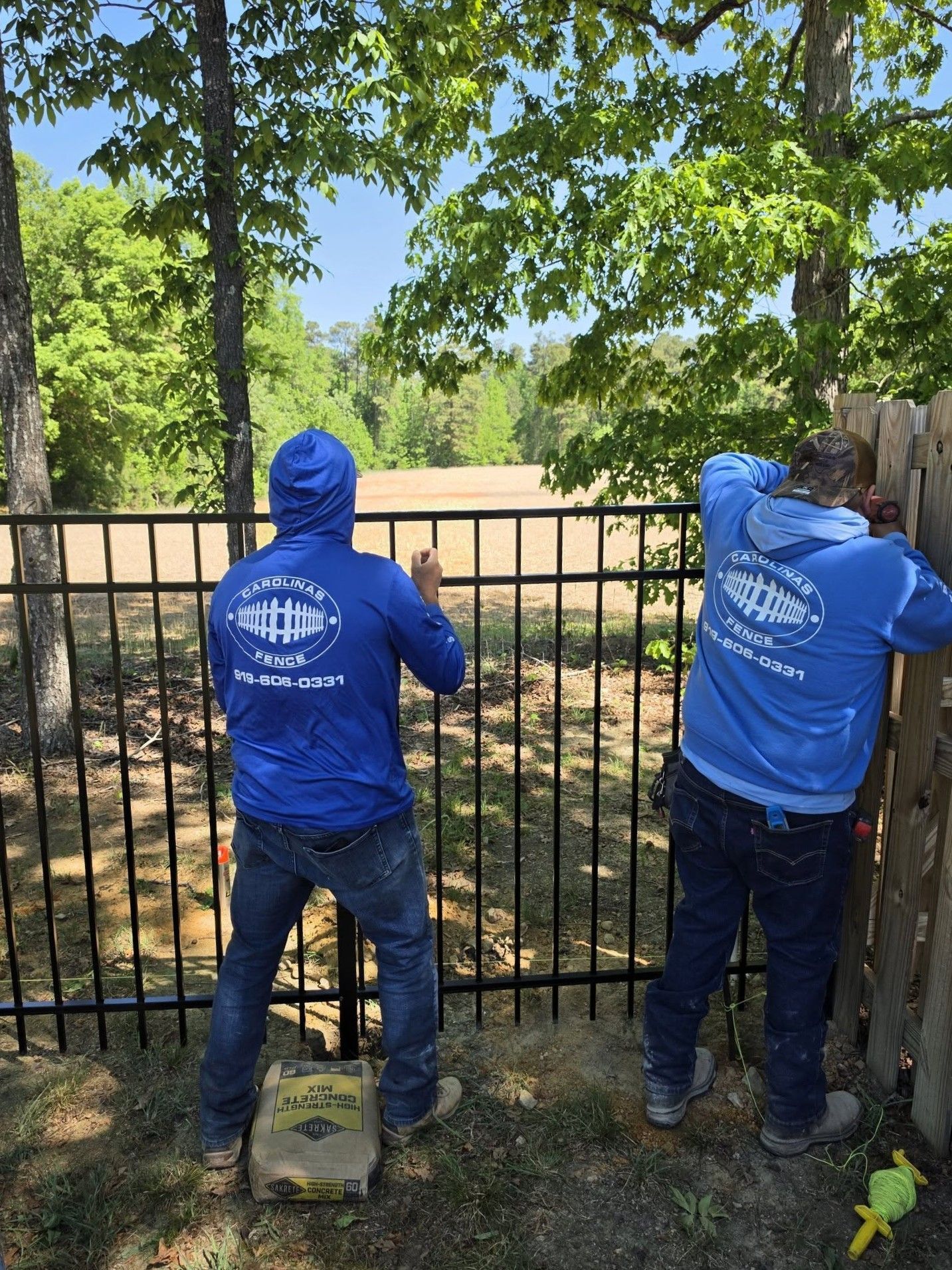 Two workers in blue hoodies install a black metal fence beside a tree-lined yard.