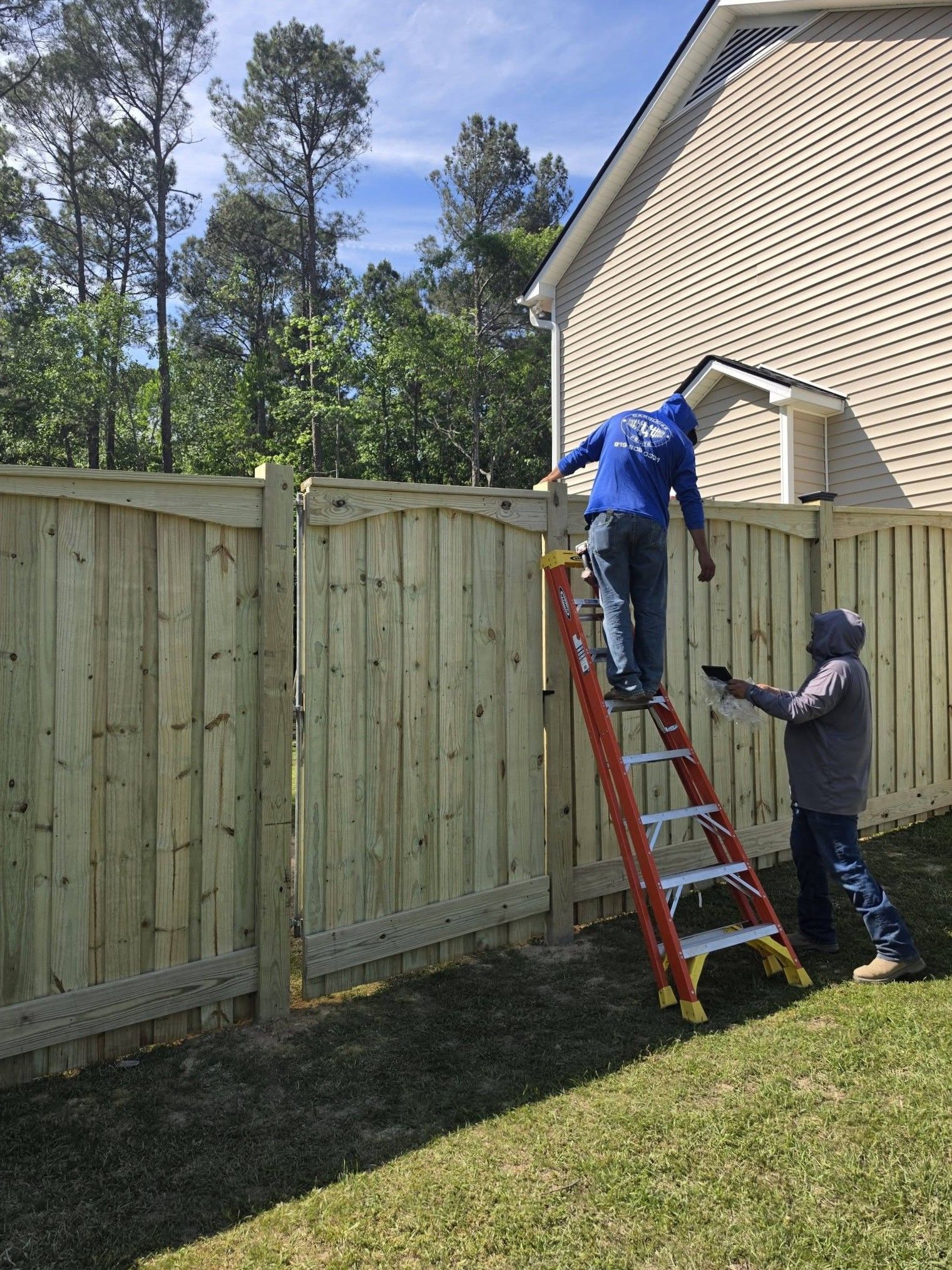 Two workers on a ladder installing a wooden fence beside a house on a sunny day