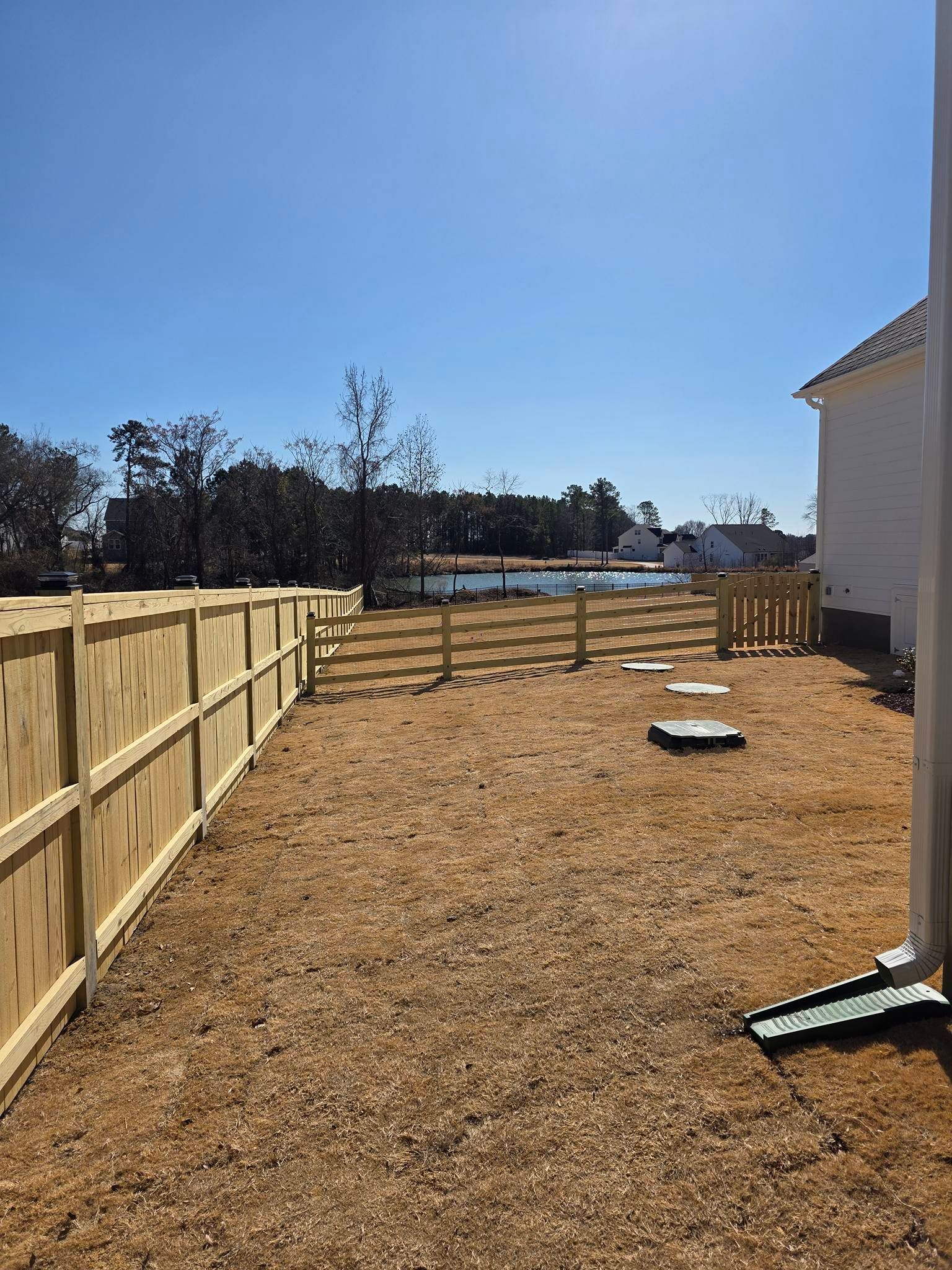 Long narrow backyard with a wooden fence, house on right, and wood chips under a clear blue sky.