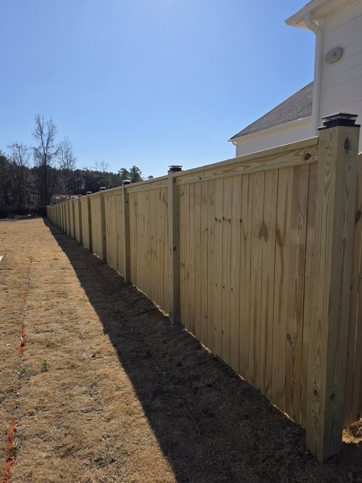 Wooden privacy fence along a house beside a dry yard under a clear blue sky