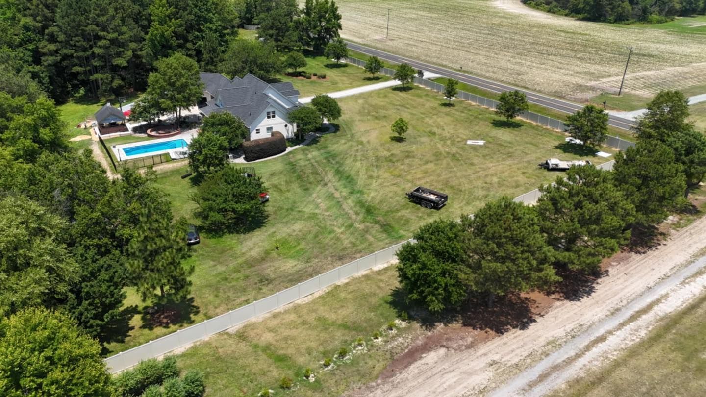 Aerial view of a rural house with a swimming pool, lawn, trees, and a gravel driveway beside a field.