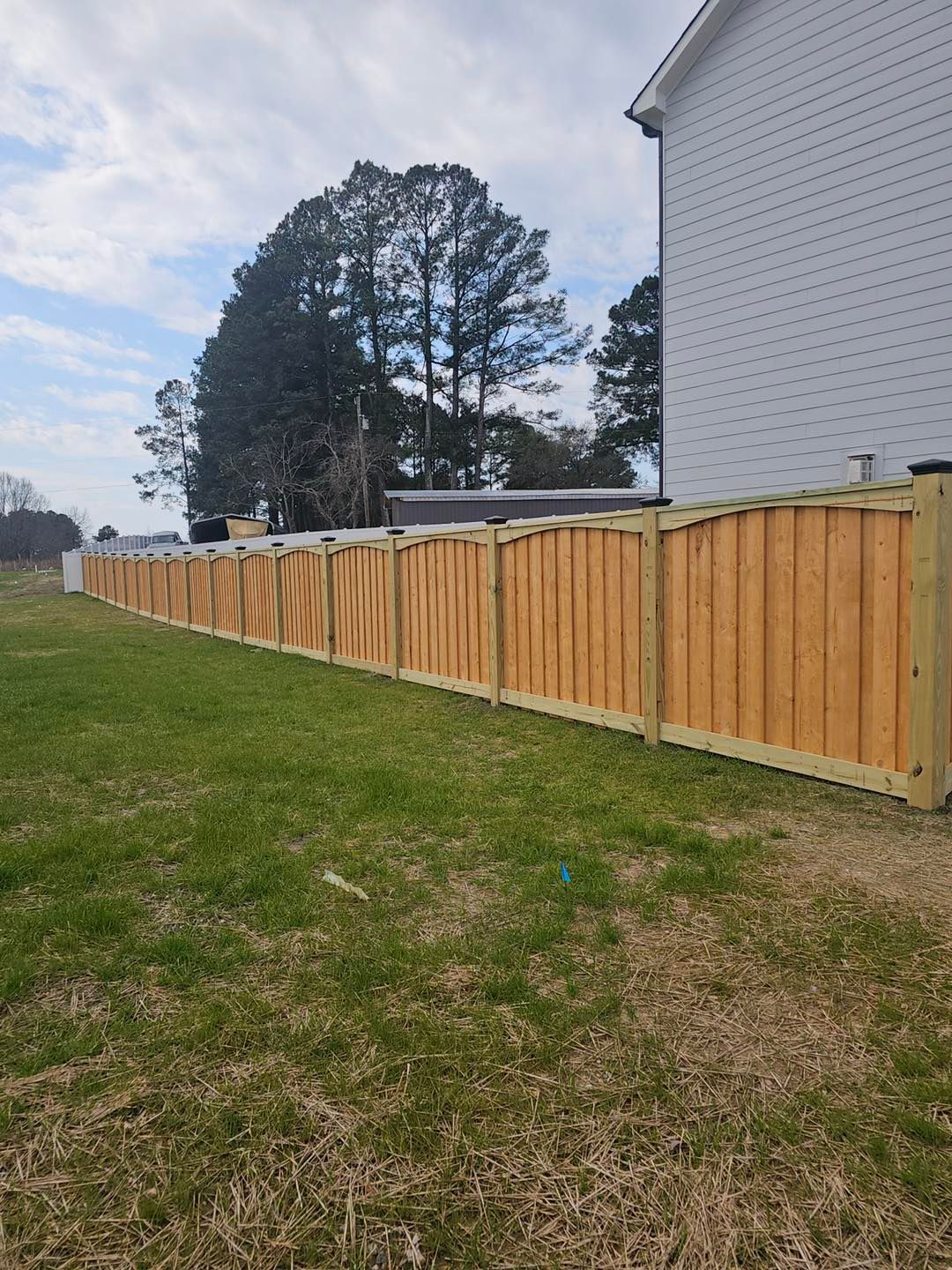 Wooden privacy fence along a grassy yard beside a white brick building and tall trees under a cloudy sky