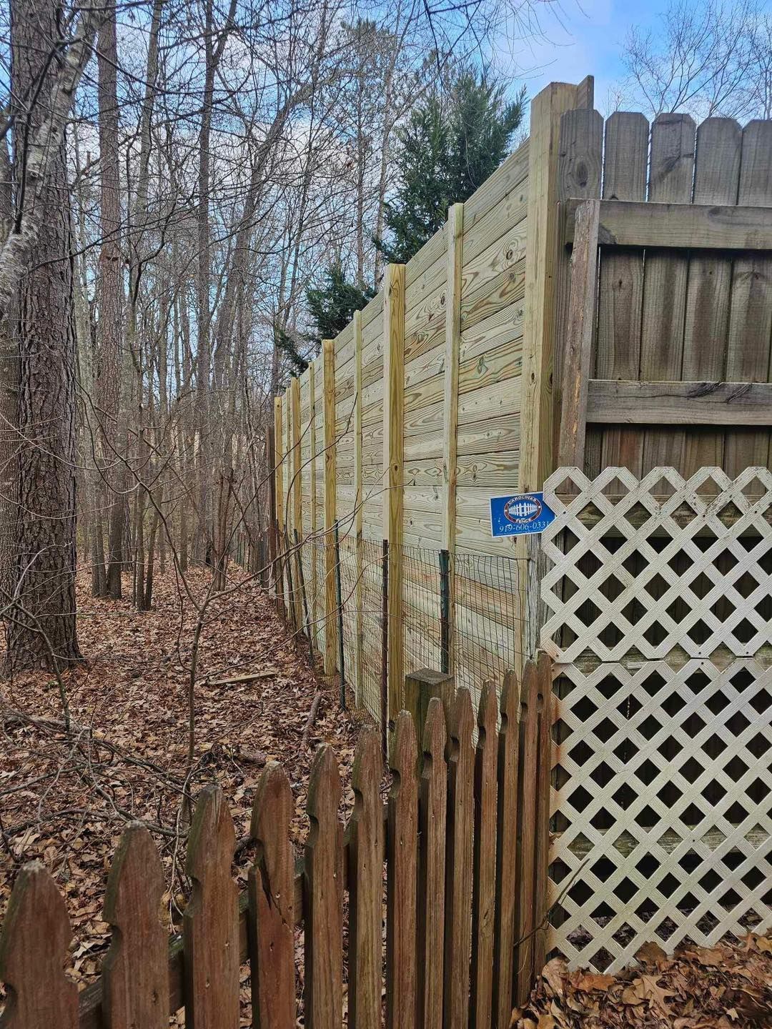 Wooden fence beside a wooded area, with lattice panels and a small blue sign.