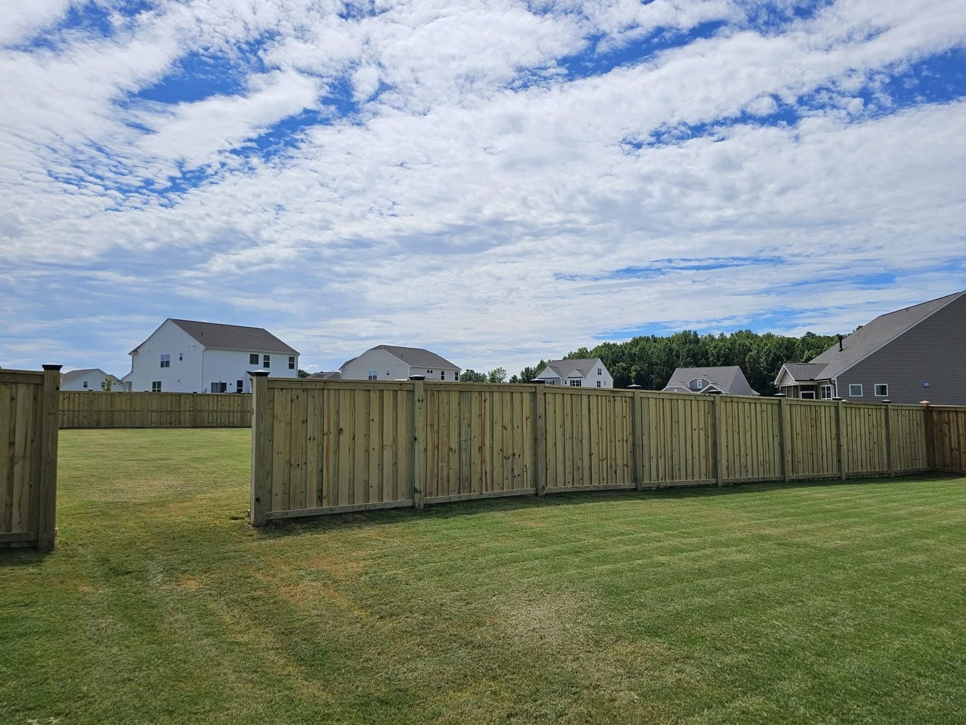 Wide grassy backyard with wooden fence, houses in the distance, and a cloudy blue sky