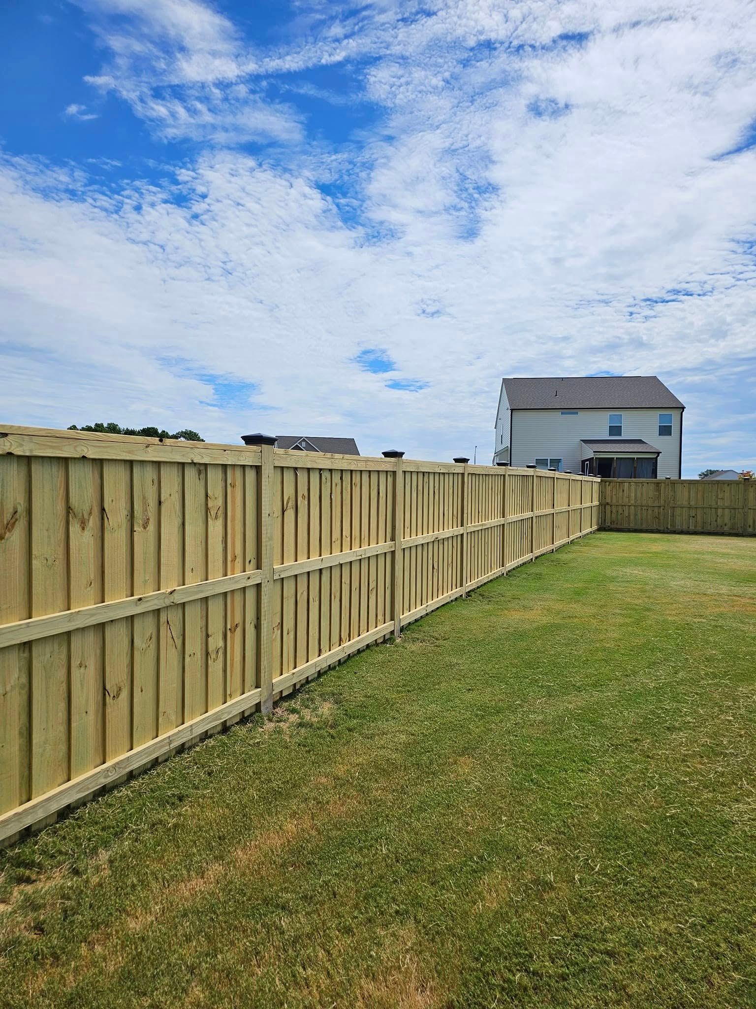 Backyard with a wooden fence, grassy lawn, and a house under a partly cloudy blue sky.