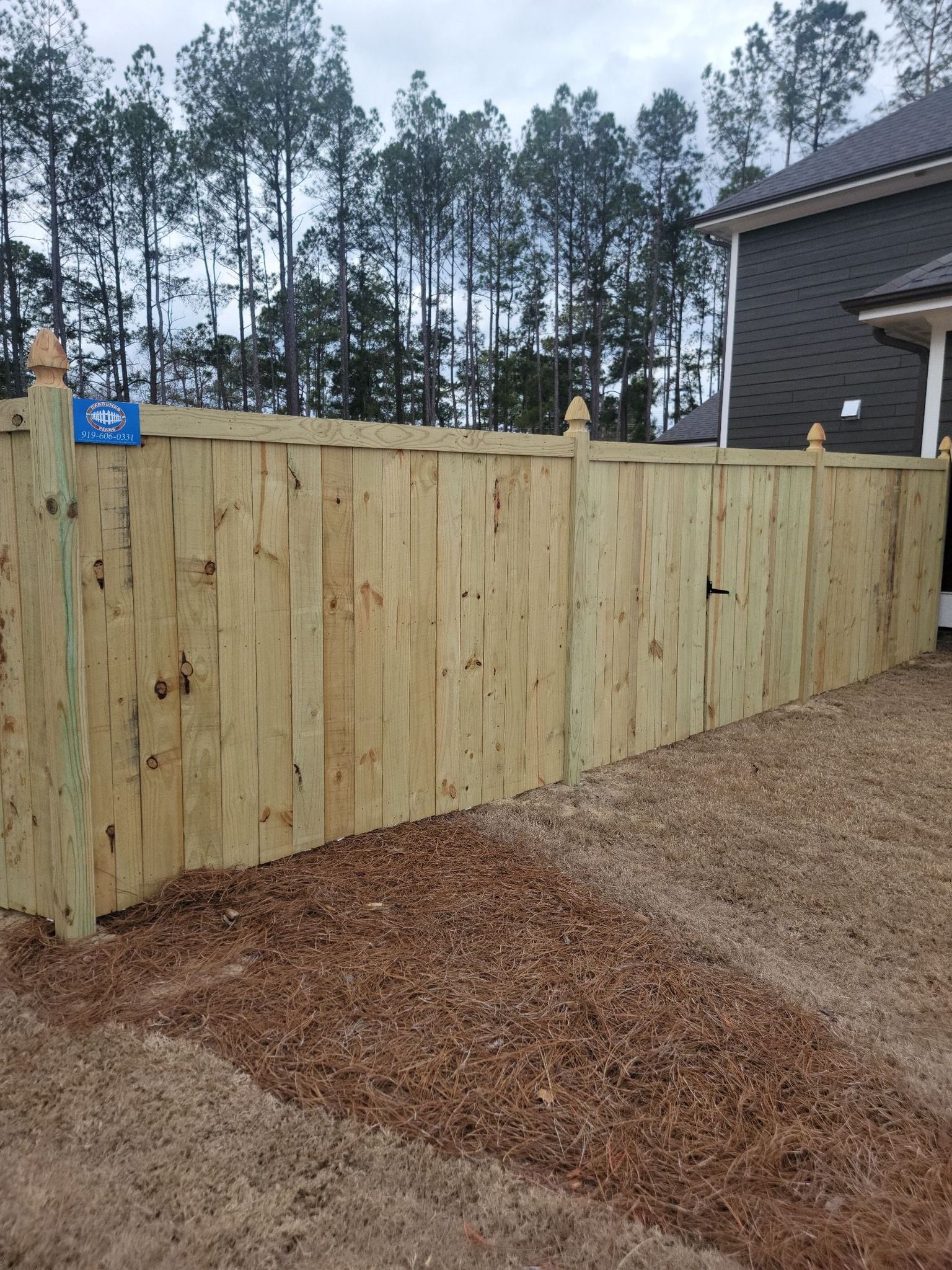 Wooden privacy fence beside a house, with trees in the background and mulch-covered ground in front.
