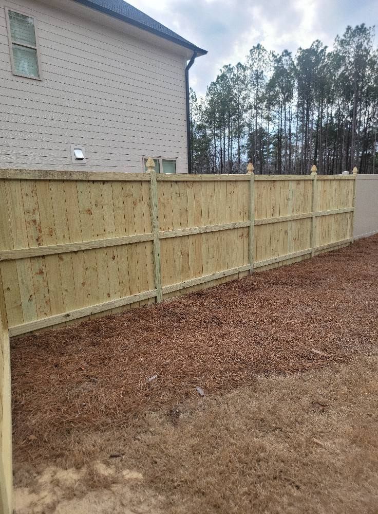 Wooden privacy fence beside a beige house, bordering a bare dirt yard with trees in the background.