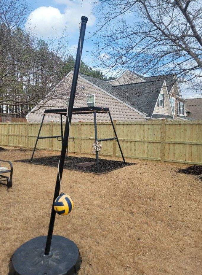 Backyard volleyball setup with net, pole, and ball on a concrete base, beside a wooden fence