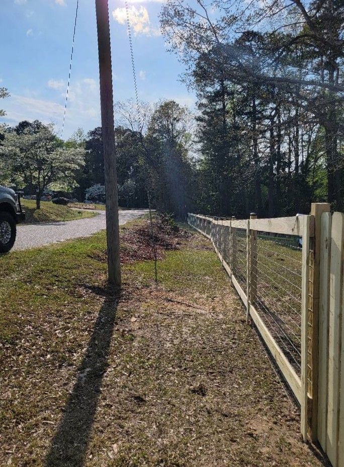 Sunlit dirt road beside a wire fence, utility pole, and trees under a clear sky