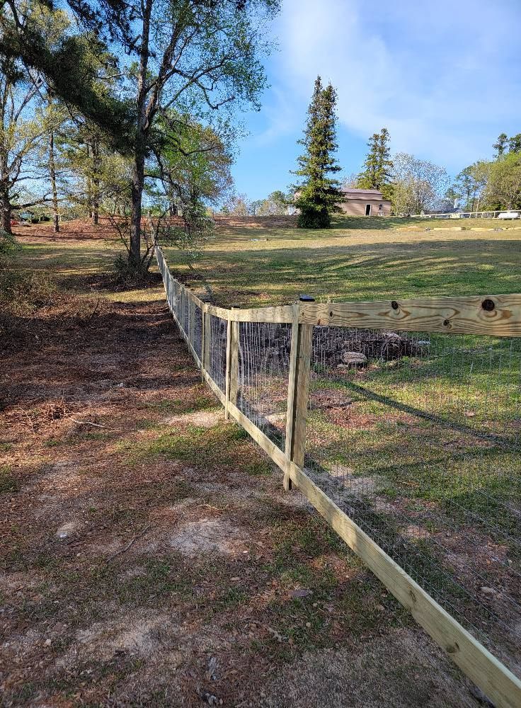 Wooden fence beside a grassy field with trees and a small house under a blue sky