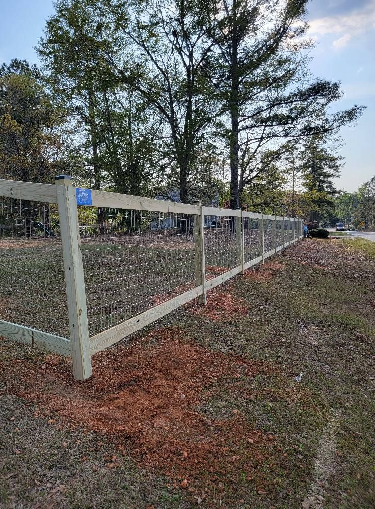 White wooden fence along a gravel road with trees and a parked black vehicle in the background.