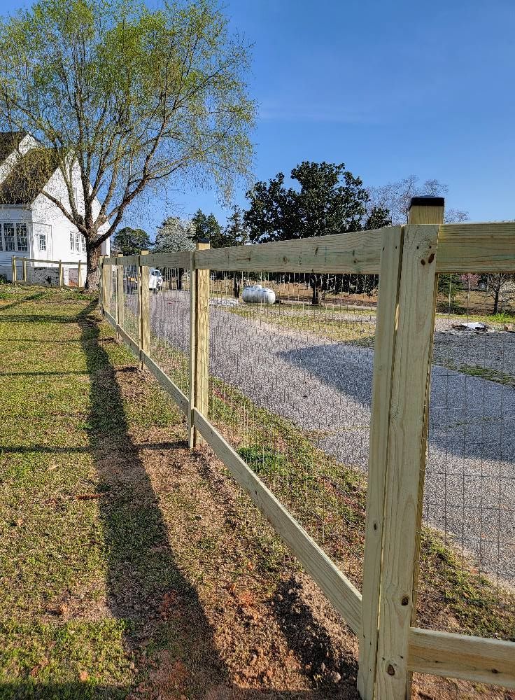 Wooden fence beside a gravel driveway, with a house and tree under a clear blue sky.