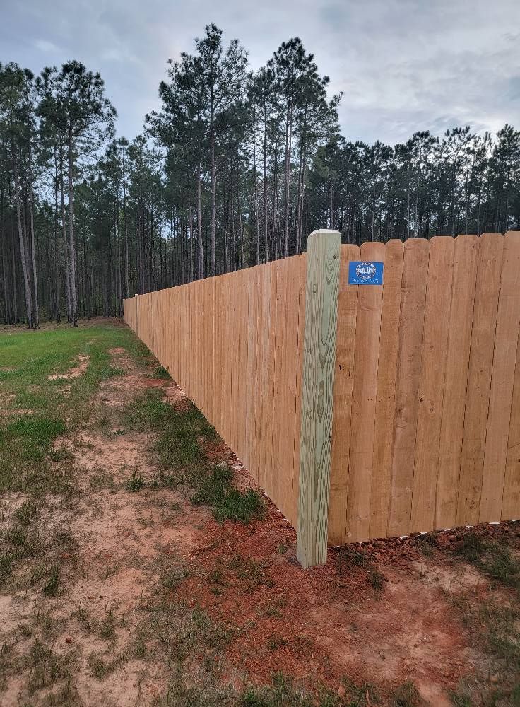 Wooden privacy fence with a blue sign along a dirt path, bordered by pine trees under a cloudy sky