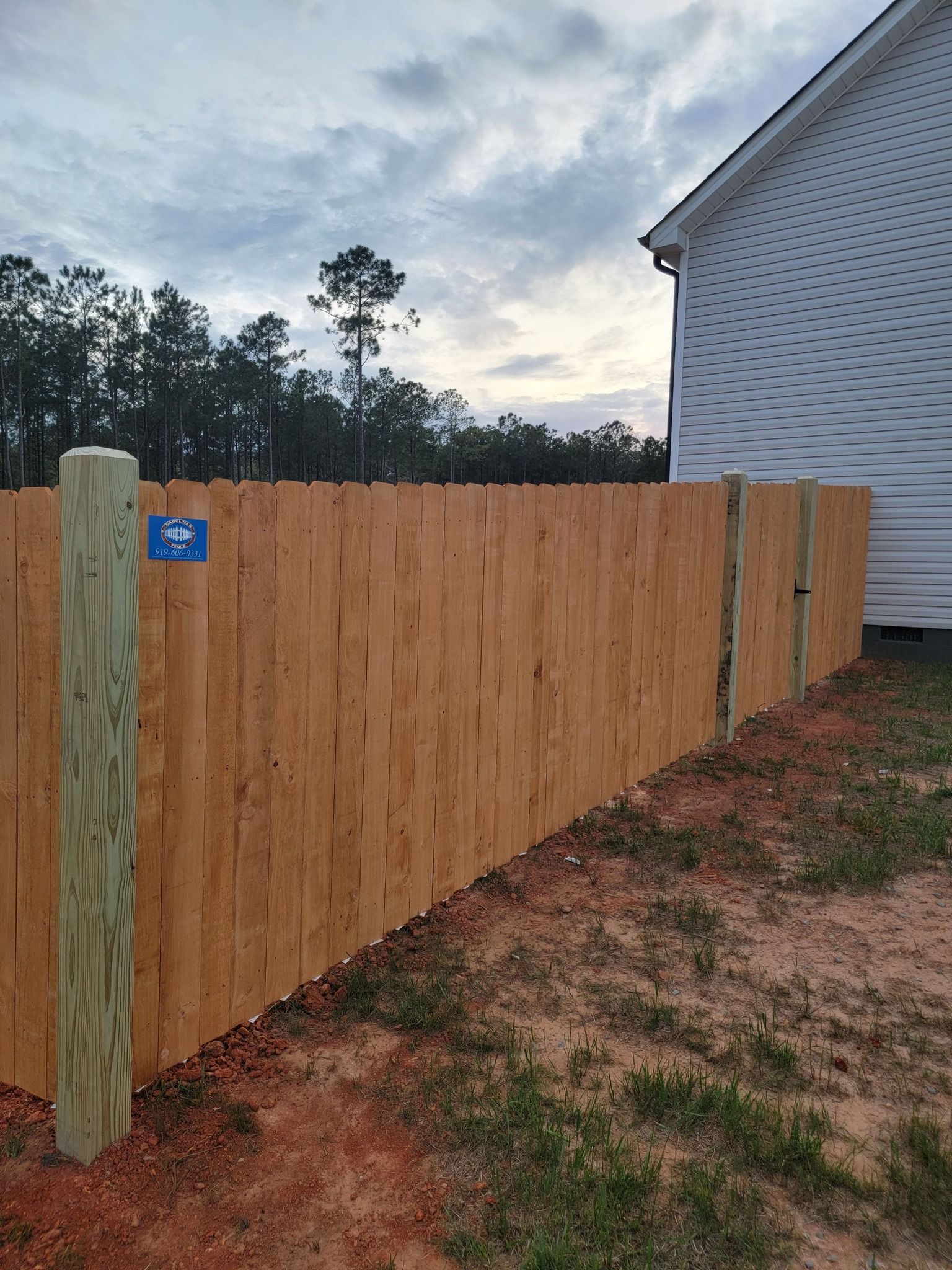 Wooden privacy fence beside a house, with a blue sign and trees in the background.