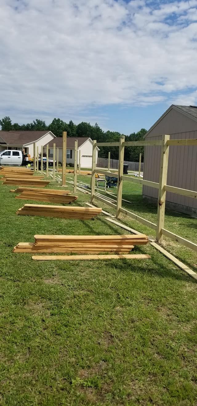 Long wooden beams laid on grass beside a fence, with houses and a partly cloudy sky nearby.