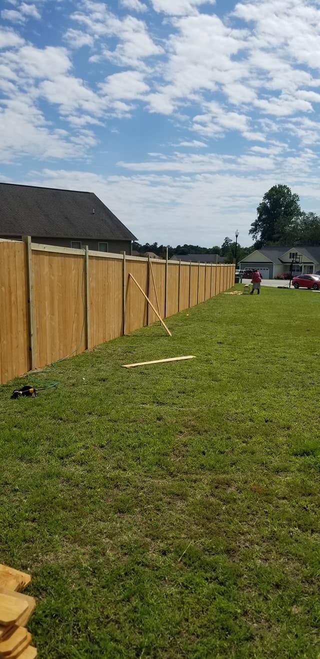 Wooden fence beside a grassy yard under a partly cloudy sky, with a house and trees in the distance