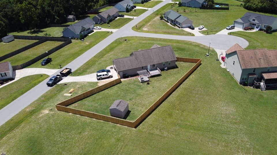 Aerial view of a suburban house with a fenced yard, driveway, parked car, and nearby homes on a green lawn.