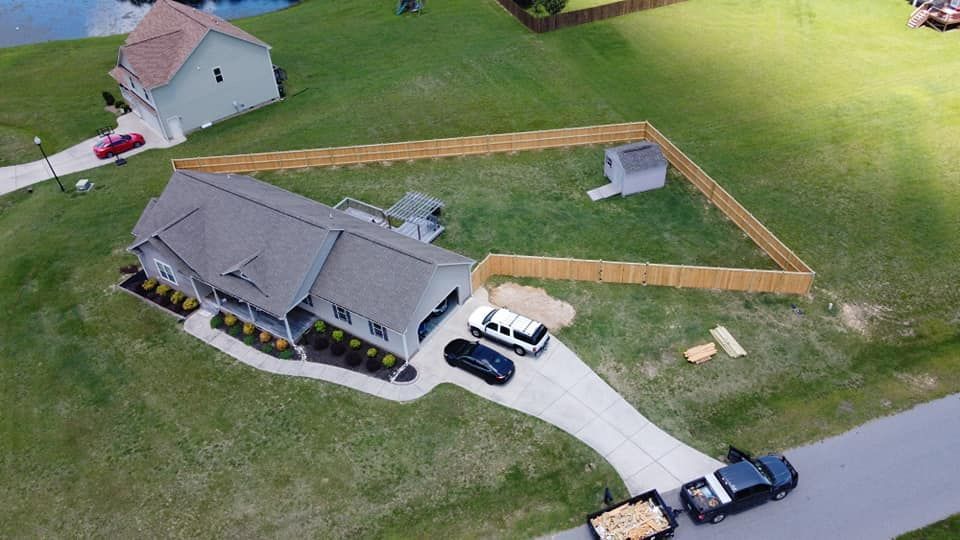 Aerial view of a house with a driveway, parked vehicles, and fenced grassy yard in a suburban neighborhood