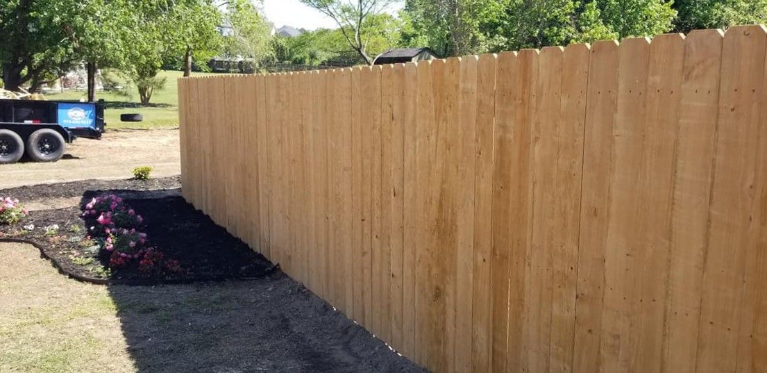 Long wooden privacy fence beside a mulched flower bed and dirt driveway under trees