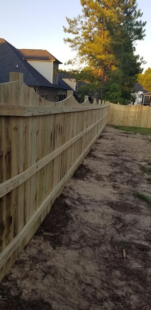 Wooden fence beside a dirt path, with a house and tall tree in the background at sunset.