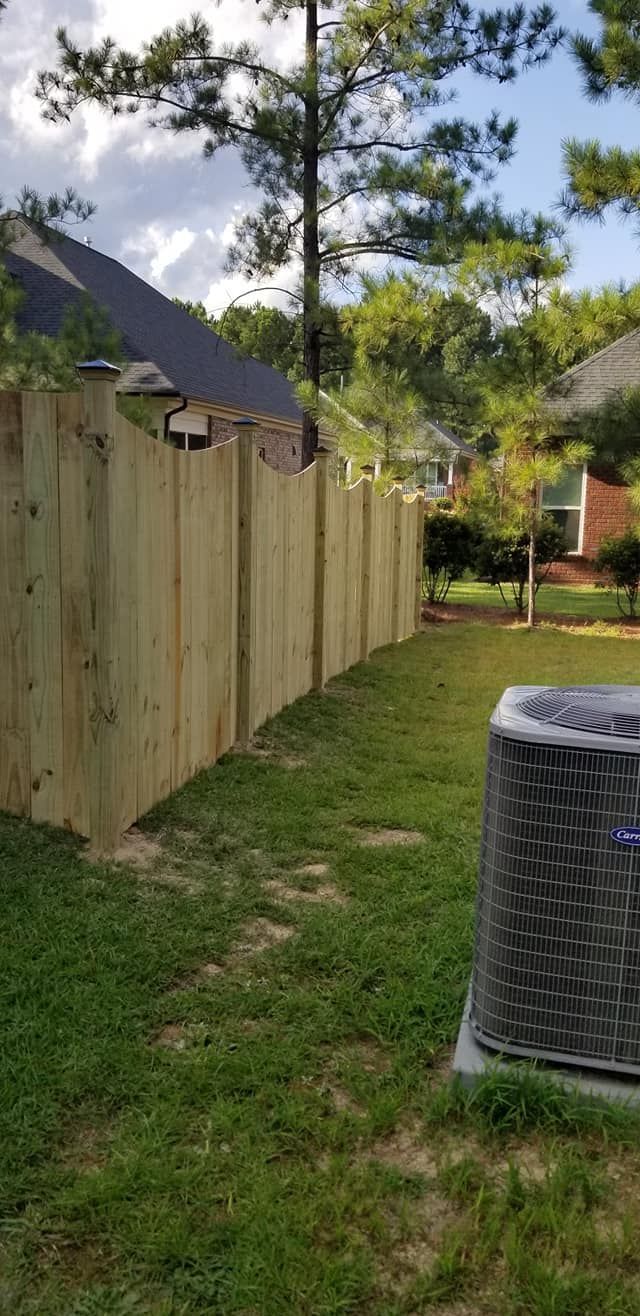 Wooden fence beside a grassy yard with trees and a house roof, plus an air conditioner unit in the foreground