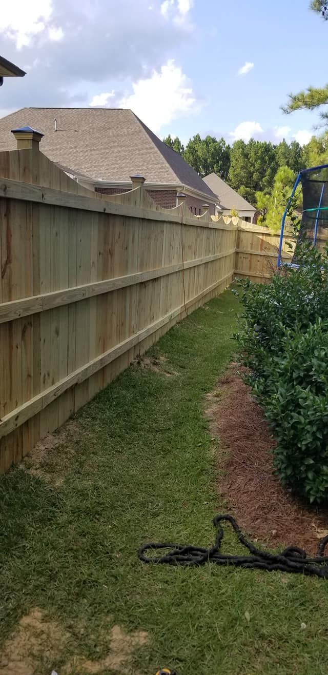 Narrow grassy side yard beside a wooden fence, with houses and trees in the background.