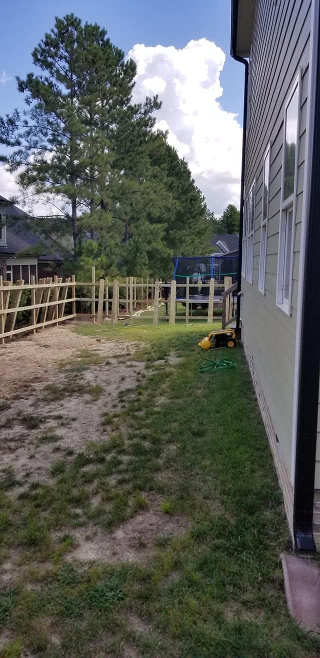 Outdoor yard beside a gray house, with a wooden fence, dirt path, and tall trees under a cloudy sky