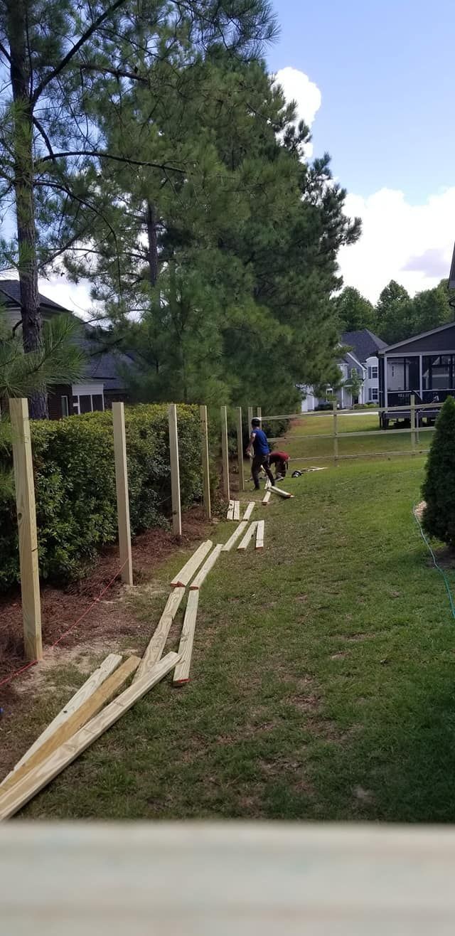Workers installing a wooden fence along a grassy yard beside tall trees and houses