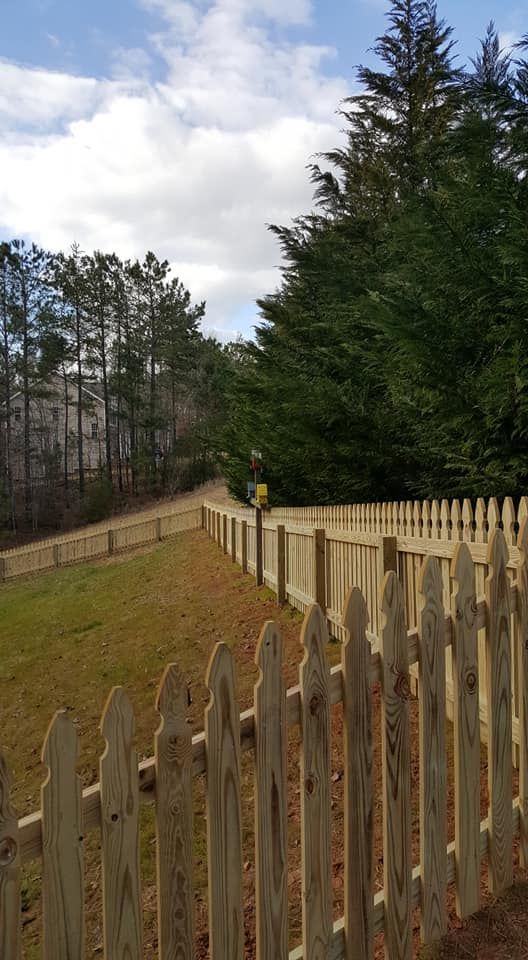 Wooden fence along a grassy path beside tall evergreens under a cloudy sky