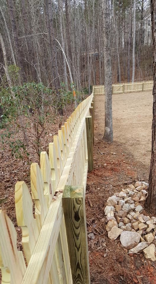 Wooden fence along a dirt path in a wooded area, with a stone border and tree trunk nearby.