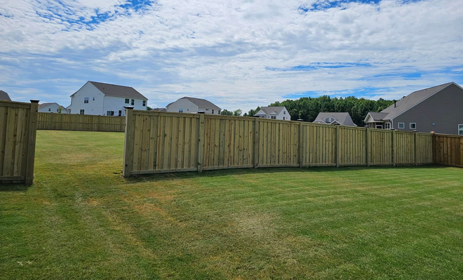 Grassy backyard with a wooden fence, suburban houses, and a partly cloudy sky