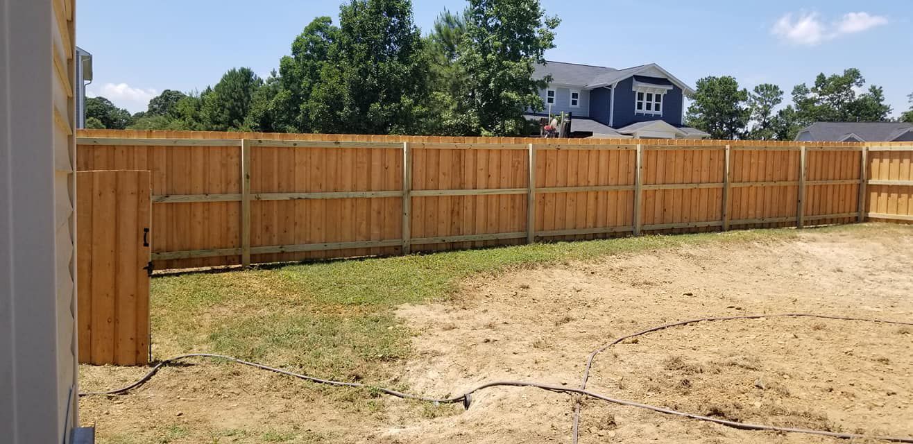 Backyard with a wooden privacy fence, dirt ground, and green trees under a bright sky