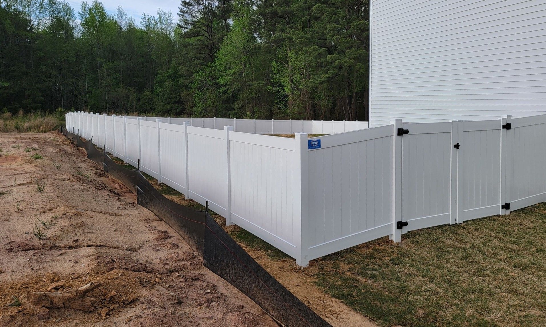 White vinyl privacy fence installed along a house and dirt lot, with wooded trees in the background.