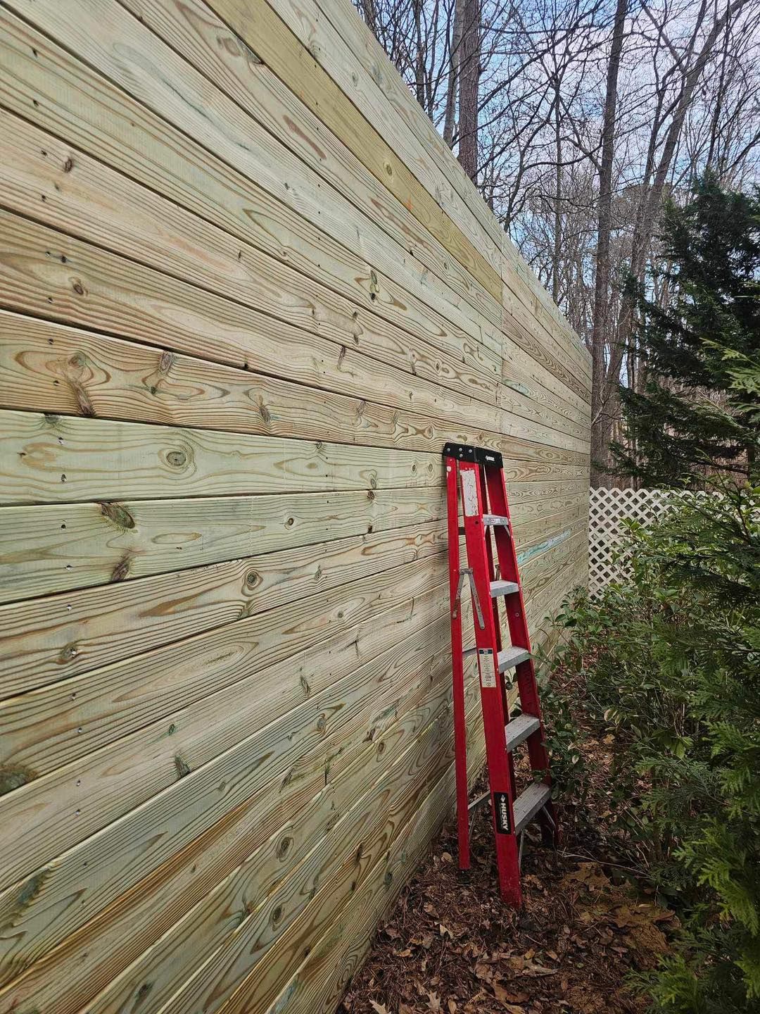 Red ladder leaning against a wooden exterior wall beside bushes and trees.