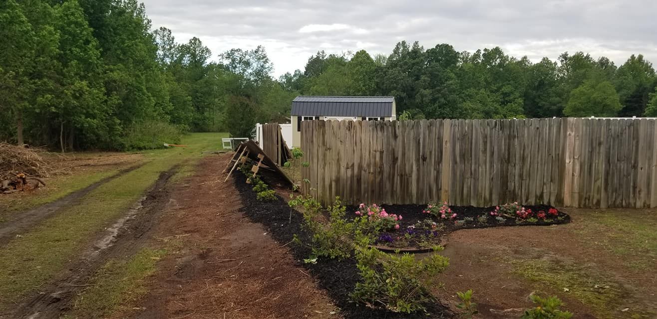Dirt driveway beside a wooden fence and flower bed, with trees and a small shed in the background.