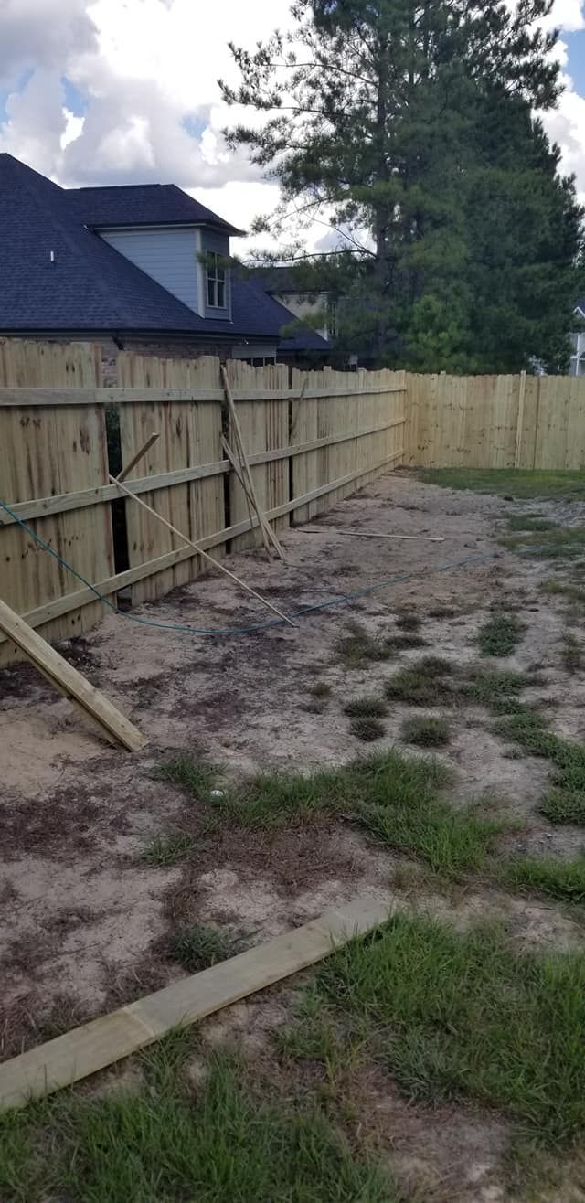 Dirt yard beside a wooden privacy fence and house, with patches of grass and trees overhead
