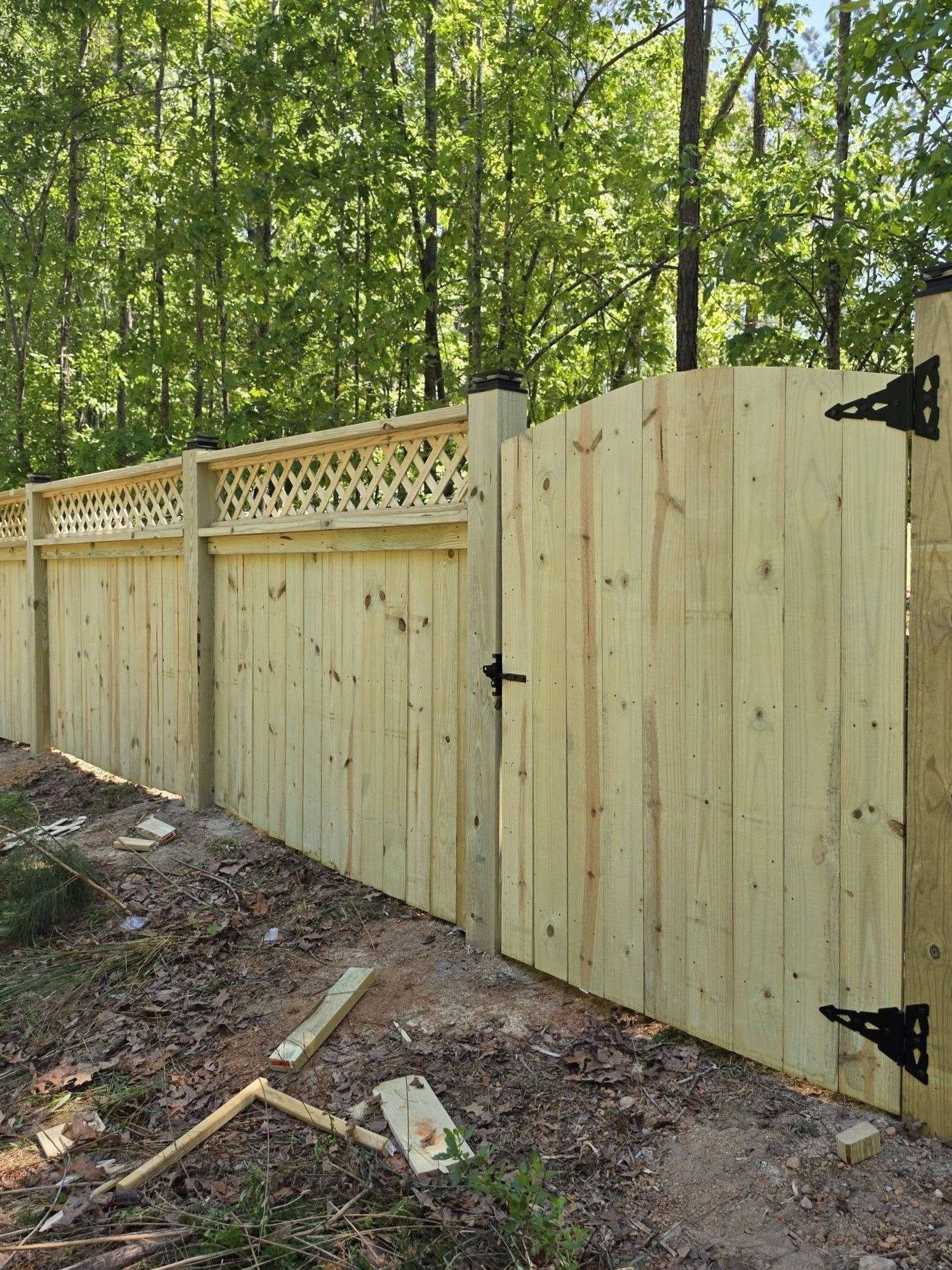 Wooden privacy fence and gate along a wooded yard, with lattice top and black hinges