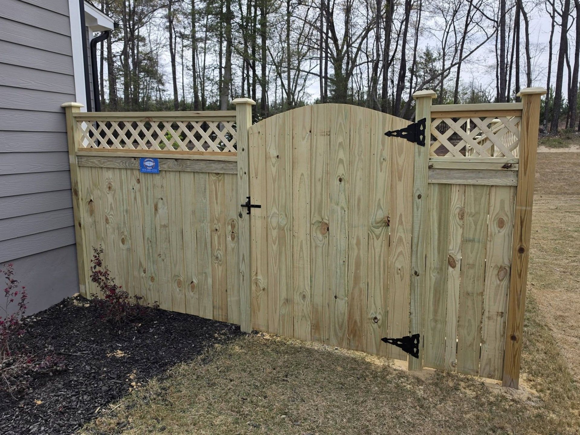Wooden garden gate and fence with lattice tops beside a house, with trees in the background.