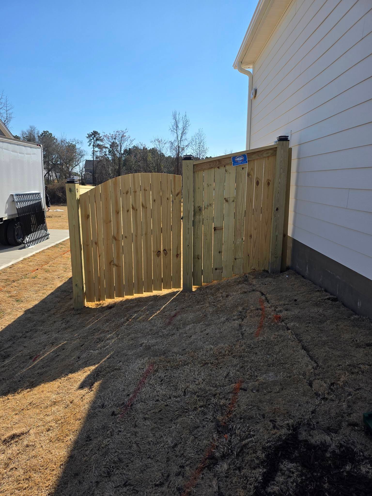 Wooden gate beside a white house on a sunny dirt path, with another white building in the background.
