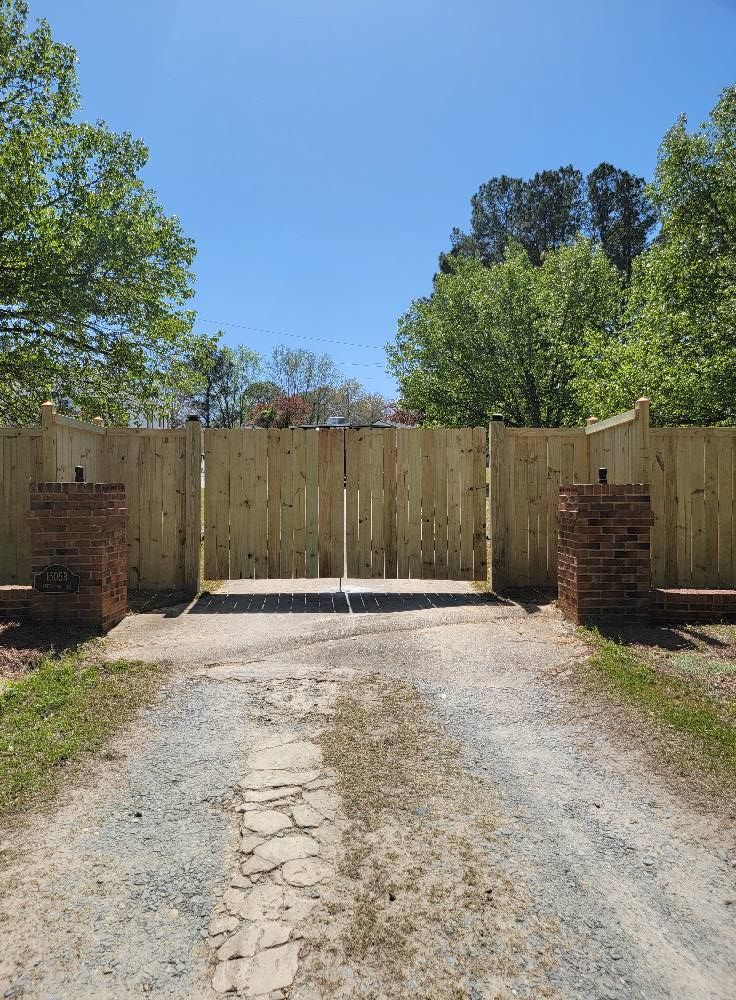 Gravel driveway leading to a closed wooden gate between brick pillars under a clear blue sky.