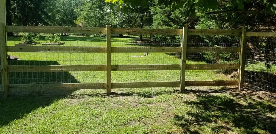 Wooden farm gate with wire mesh in a grassy field, backed by trees and sunlight