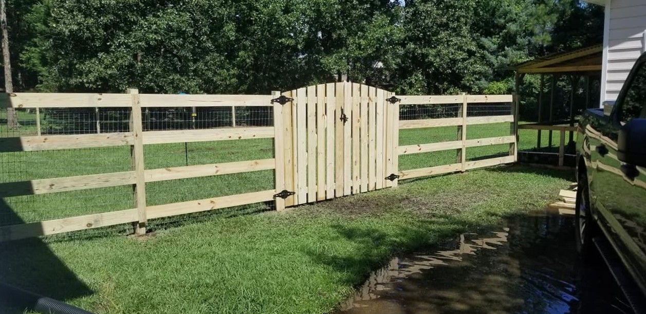 Wooden fence with a central gate across a grassy yard beside a driveway and trees