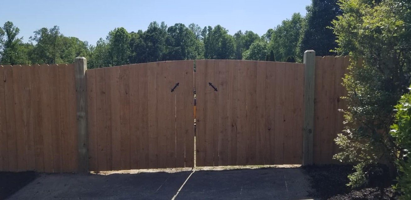 Closed wooden double gate with a central latch in a fenced yard, trees and shrubs behind it.