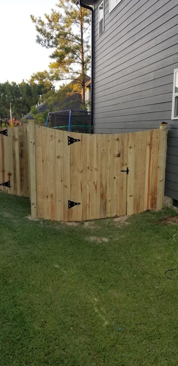 Wooden fenced enclosure beside a gray house, with grass and trees in the yard.