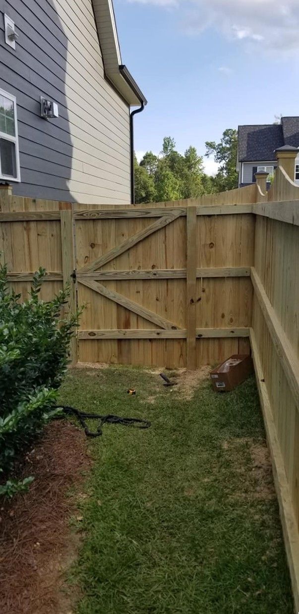 Narrow backyard with grass, wooden fence gate, and side of a house under a blue sky