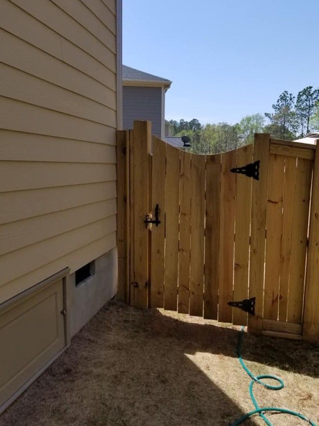 Wooden gate beside a beige house wall, with black hinges and a green hose on the dirt ground.
